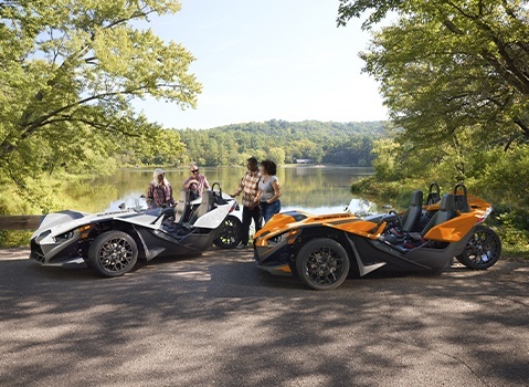 Four people standing beside a white and a orange Polaris Slingshot roadsters on a tree-lined lakeshore.