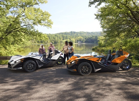 Four people standing beside a white and a orange Polaris Slingshot roadsters on a tree-lined lakeshore.