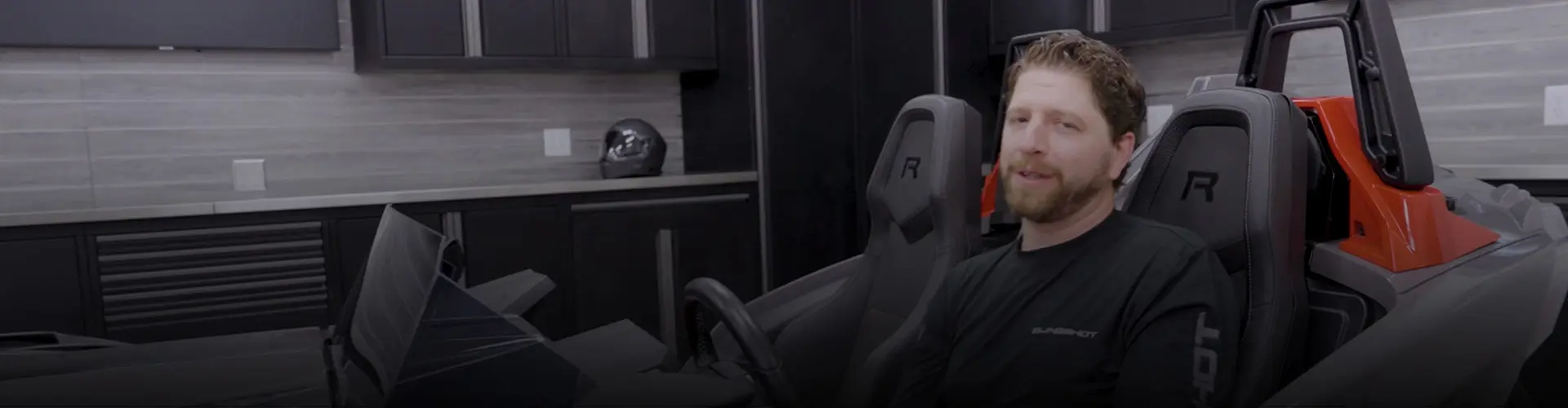Jake Stock seated inside a Polaris Slingshot Roadster in a residential garage, with branded seats, a helmet, and storage cabinets visible nearby.