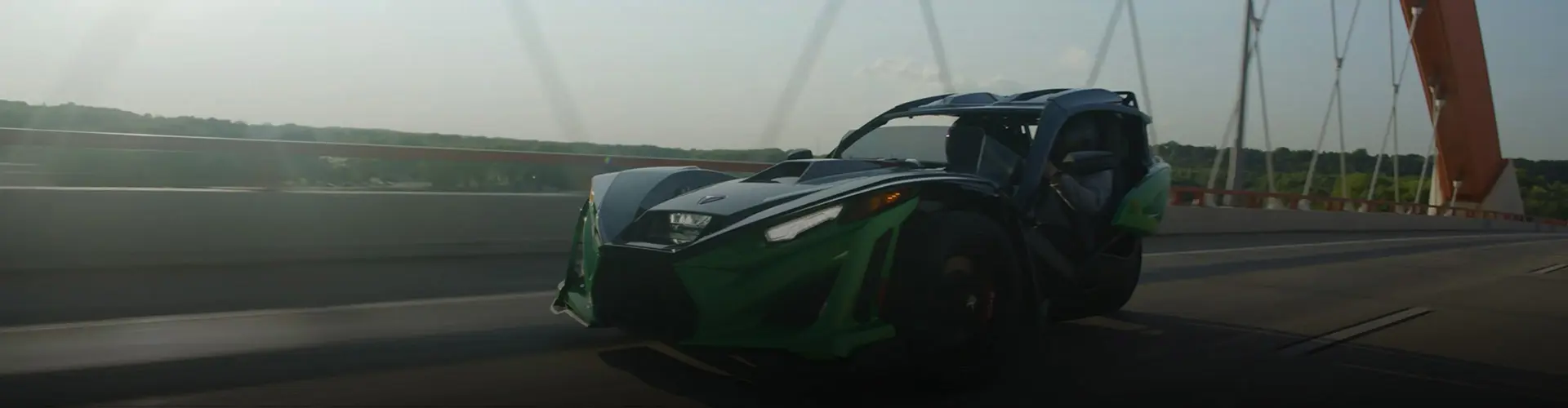 Polaris Slingshot driving across a suspension bridge, captured in motion with green bodywork and forested scenery in the background.