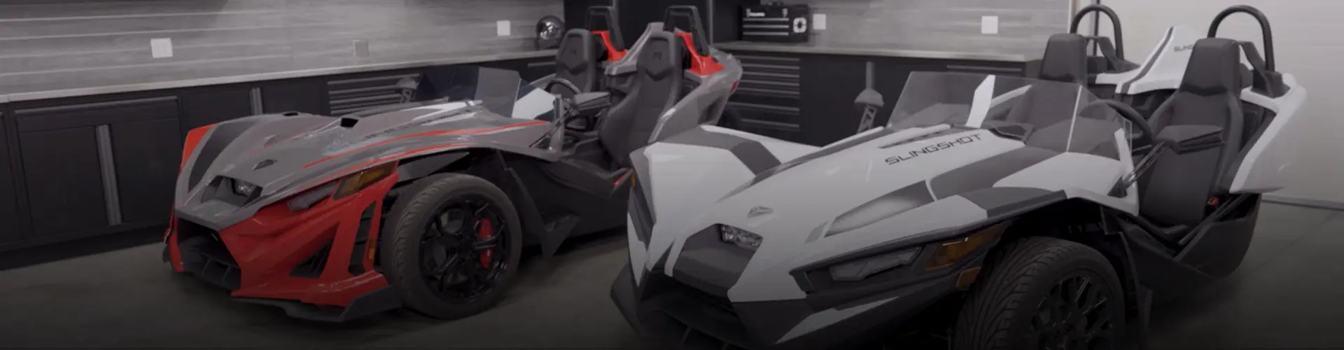 Two Polaris Slingshot three-wheeled roadsters parked side by side in a modern garage, featuring red, gray and white bodywork.