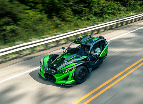 Polaris Slingshot driving along a winding two-lane road, captured from above with green bodywork and surrounding forest blurred by motion.