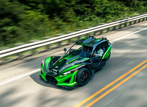 Polaris Slingshot driving along a winding two-lane road, captured from above with green bodywork and surrounding forest blurred by motion.