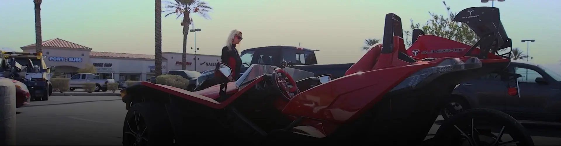 Red Polaris Slingshot roadster parked in a shopping center lot with Melody besides.