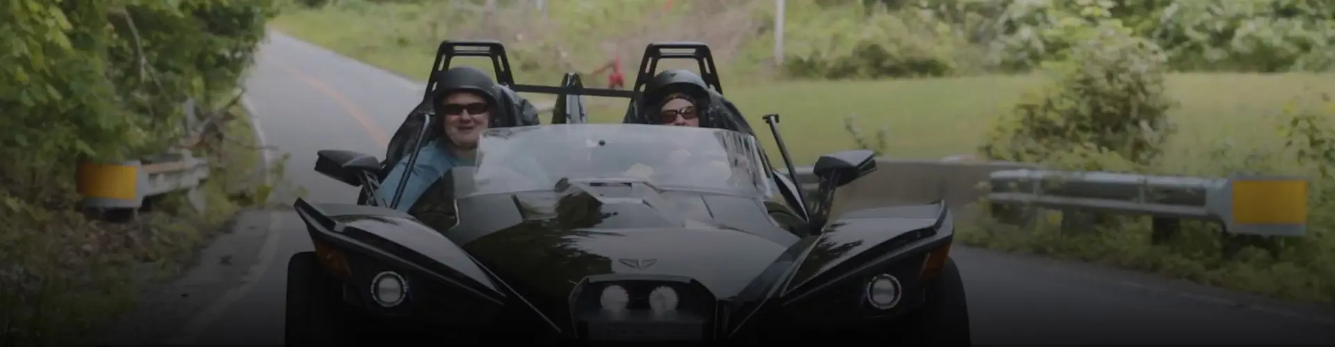Kelly and Blake wearing helmets ride a black Polaris Slingshot autocycle traveling along a tree-lined rural road.