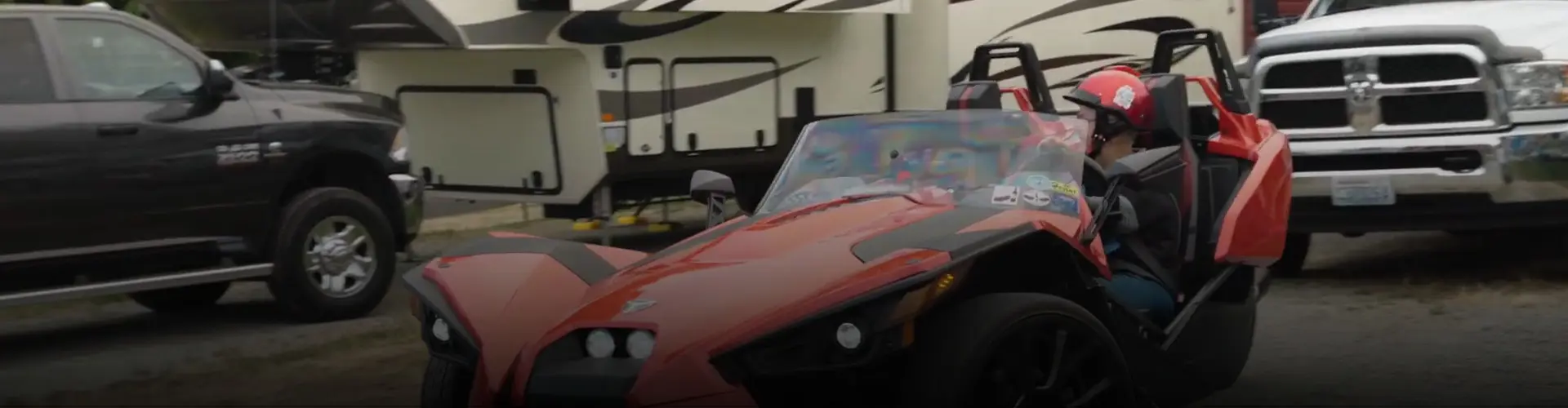 Anita wearing a helmet in a red Polaris Slingshot roadster passing trucks and RVs at a campground.