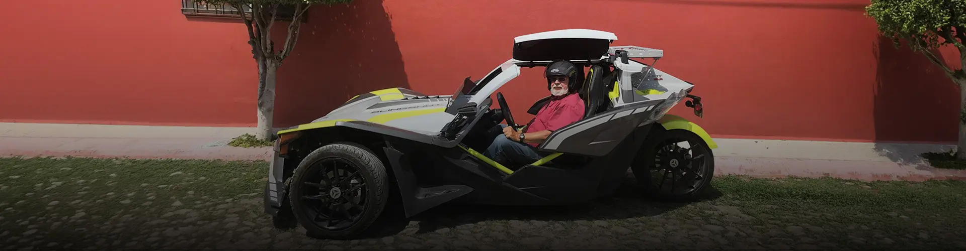 Custom gray and yellow Polaris Slingshot roadster with a roof and parked beside a red wall with Daryl in the driver seat wearing a helmet.