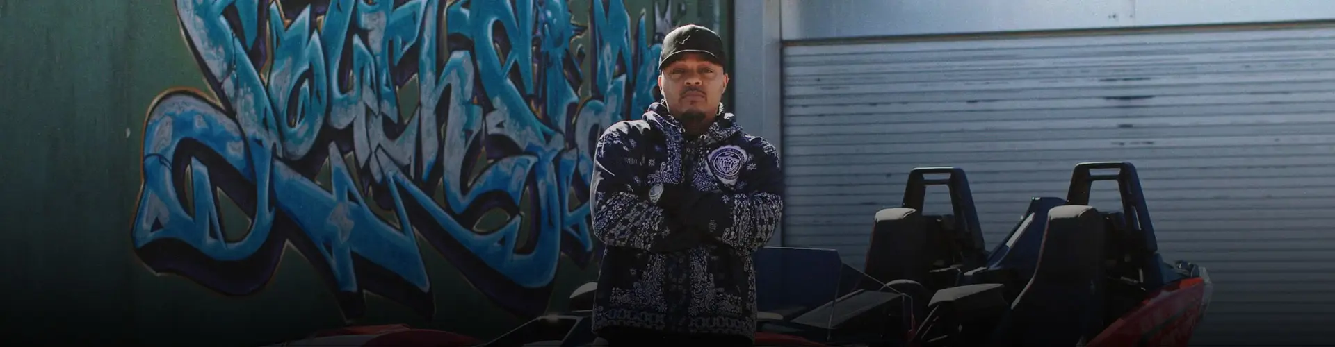 Bow Wow standing with arms crossed beside a red Polaris Slingshot roadster in front of a blue graffiti mural.