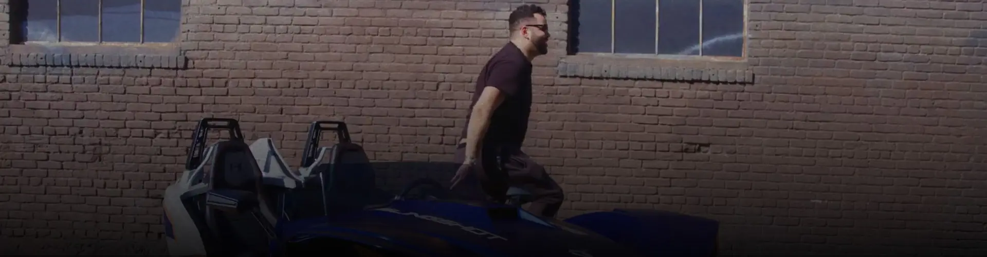 Jeremy Fall stepping out of a blue Polaris Slingshot autocycle parked beside a brick building.