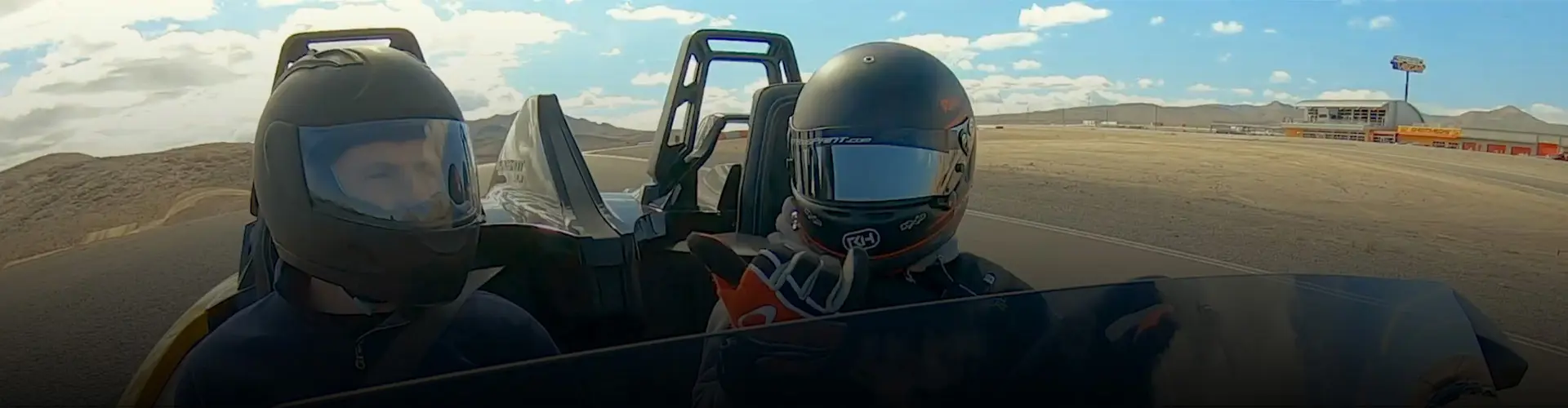 Two helmeted riders aboard a Polaris Slingshot roadster on a desert racetrack.