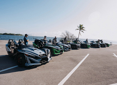 Polaris Slingshot three-wheel vehicles lined up with riders in a waterfront parking lot.