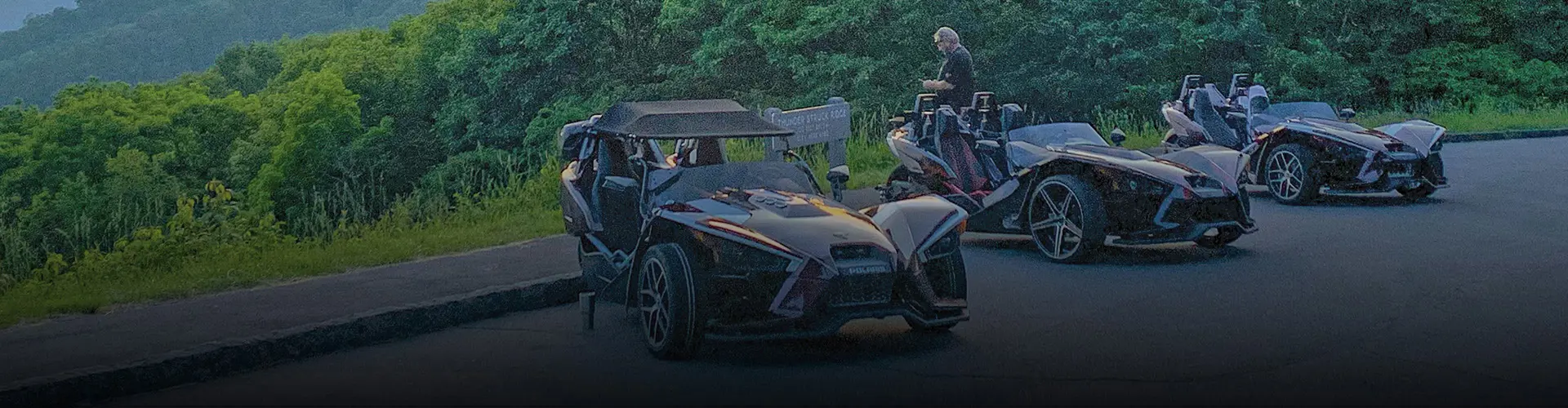 Three custom Polaris Slingshot roadsters parked at a scenic overlook surrounded by forested mountains.