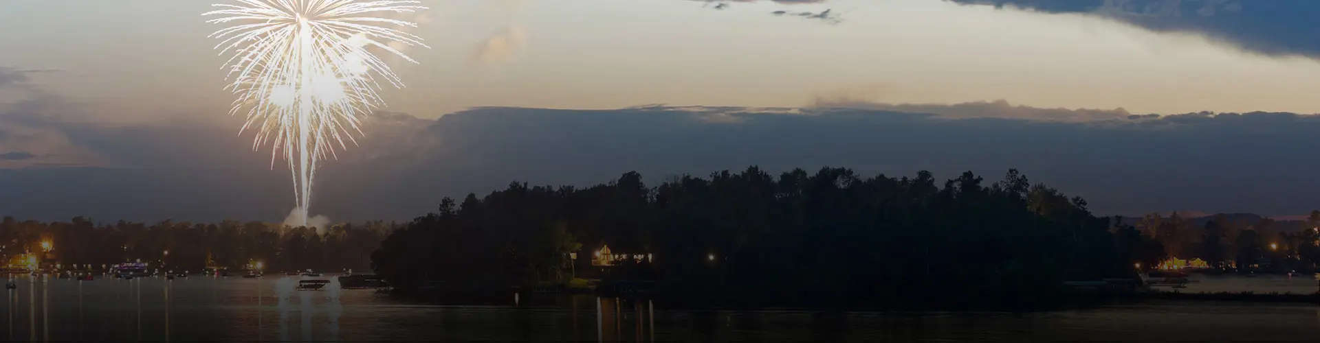 White fireworks illuminate the evening sky above a tree-lined lake with boats and shoreline lights.