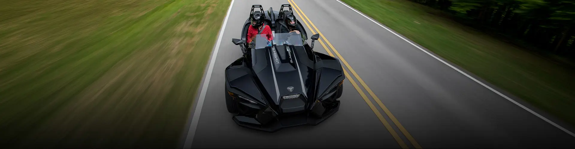 Black Polaris Slingshot three-wheel vehicle with two helmeted riders traveling on a tree-lined road.