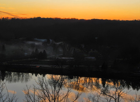 Orange sunset reflected across a calm river beneath the silhouette of a wooded landscape.