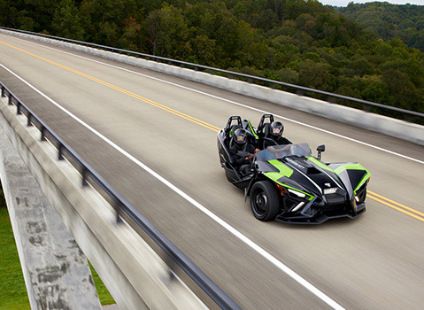 Green and black Polaris Slingshot roadster with two helmeted riders crossing a bridge above a wooded valley.