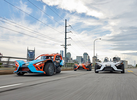 Three colorful Polaris Slingshot autocycles driving on a city highway with the Nashville skyline behind them.