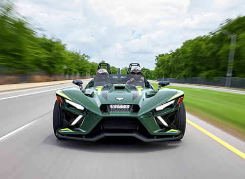 Green Polaris Slingshot autocycle with two helmeted riders traveling along a tree-lined road.