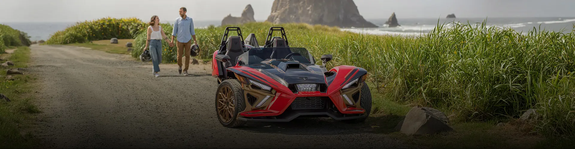 Red and black Polaris Slingshot roadster parked along a coastal path as two people carry helmets near ocean rock formations.