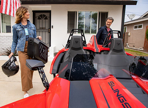 A couple prepare for a ride beside a red Polaris Slingshot three-wheel vehicle parked outside a home.