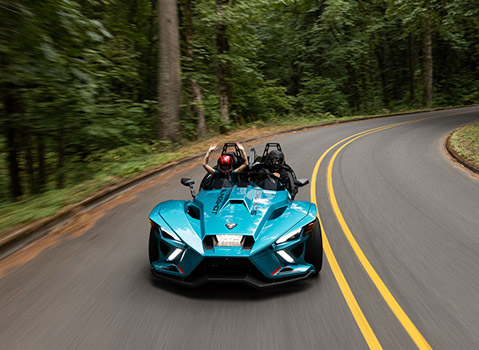 Blue Polaris Slingshot autocycle with two helmeted riders traveling along a winding forest road.