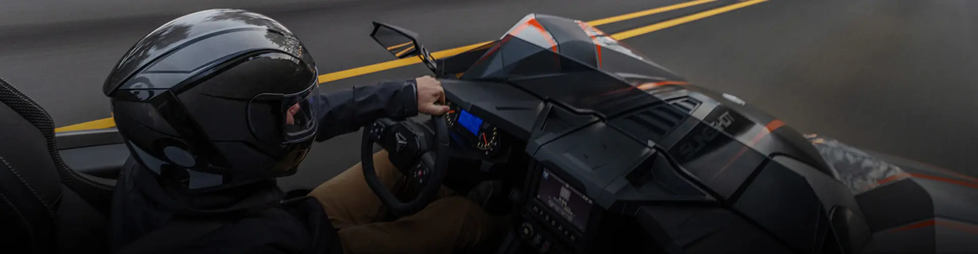 Helmeted driver operating a black and orange Polaris Slingshot roadster on a winding paved road.