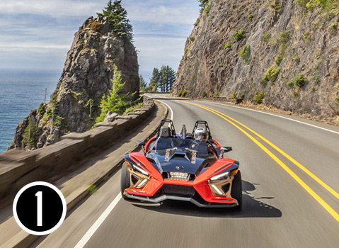 Red Polaris Slingshot roadster with a helmeted driver traveling along a winding coastal highway beside rocky cliffs.