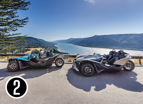 Two custom Polaris Slingshot autocycles parked at an overlook above a river surrounded by forested mountains.