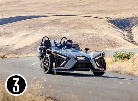 Gray Polaris Slingshot three-wheel vehicle driving on a winding road through dry rolling hills.