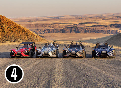 Four custom Polaris Slingshot three-wheel vehicles lined up at an overlook above a rugged desert canyon.
