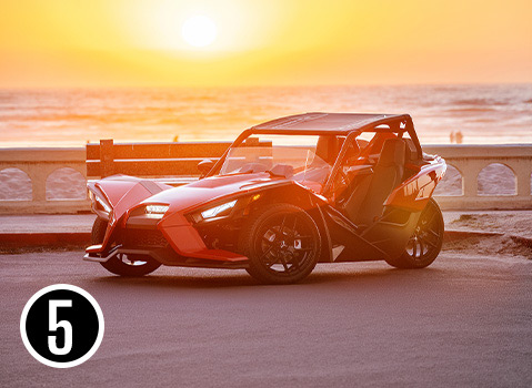 Red Polaris Slingshot roadster with a roof parked by the ocean during a golden sunset.