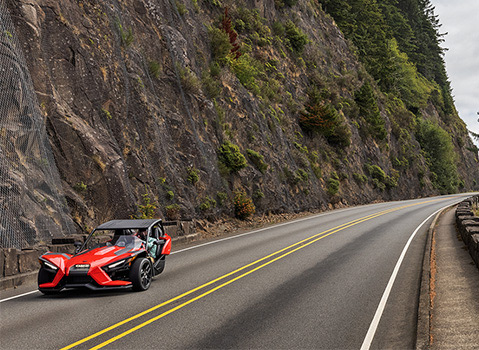Red Polaris Slingshot autocycle traveling along a winding mountain road bordered by rocky cliffs and evergreen trees.
