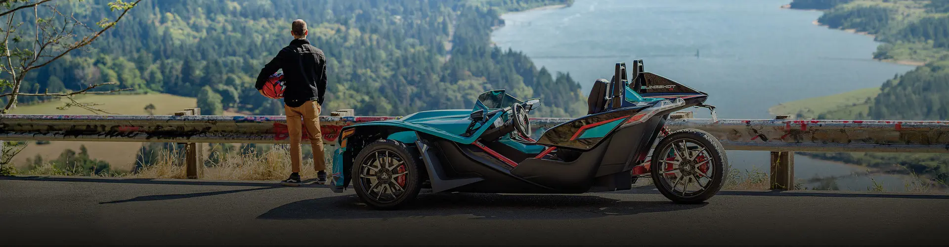 Custom teal Polaris Slingshot roadster parked at a scenic overlook as a person views the river and forested hills.
