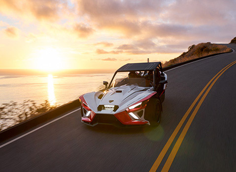 White and red Polaris Slingshot autocycle driving along a winding coastal road at sunset.