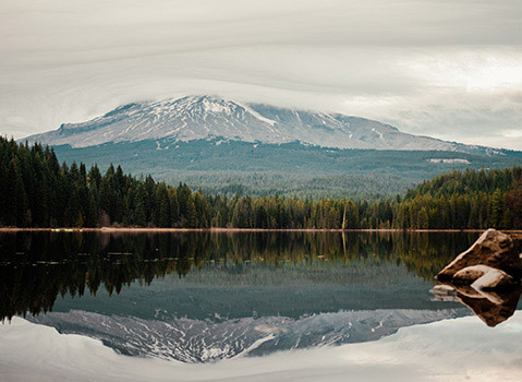Snowcapped mountain reflected in a calm lake surrounded by dense evergreen forest.