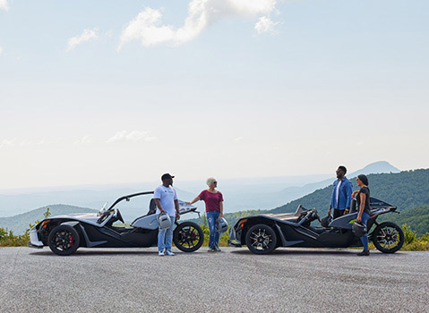 Four friends holding helmets beside two Polaris Slingshot roadsters at a scenic mountain overlook.