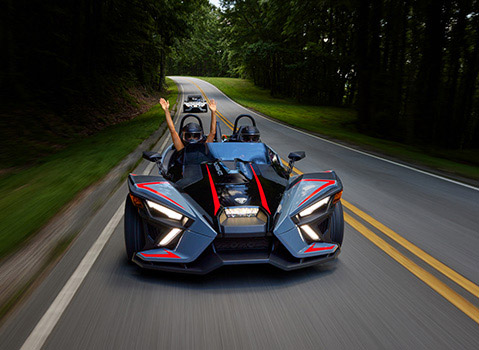 Gray Polaris Slingshot three-wheel vehicle with two helmeted riders traveling along a winding forest road.