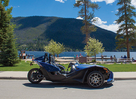 Blue Polaris Slingshot autocycle parked near a mountain lake and waterfront picnic area.