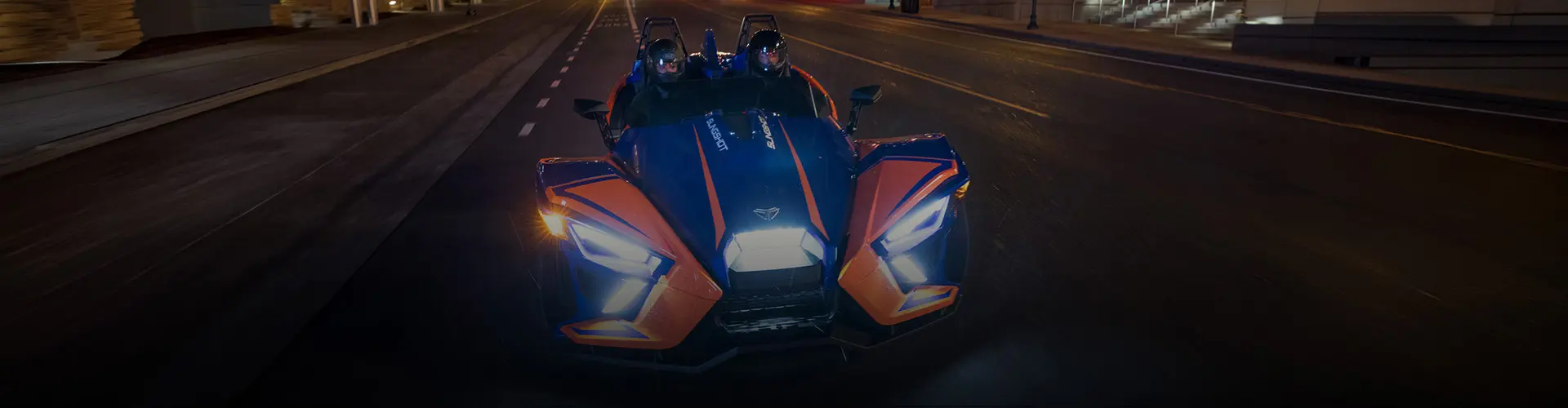 Blue and orange Polaris Slingshot autocycle with two helmeted riders driving on a city street at night.