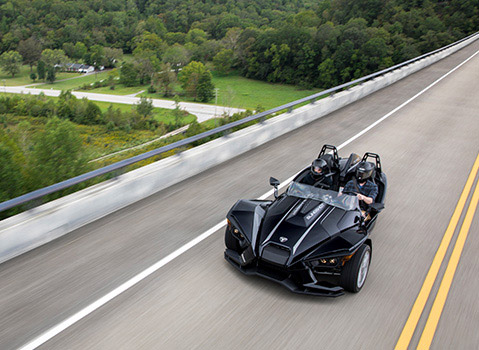 Green Polaris Slingshot roadster driving beside misty water and autumn trees in warm evening light.