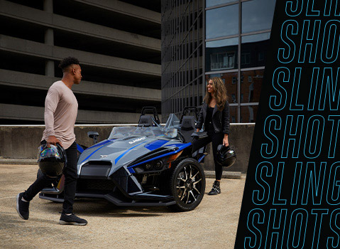 A group of friends gathered beside two Polaris Slingshot three-wheel vehicles at an outdoor venue.