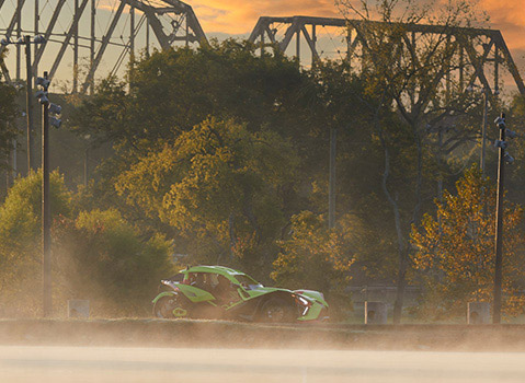 Green Polaris Slingshot roadster driving beside misty water and autumn trees in warm evening light.