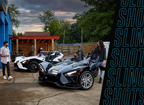 A group of friends gathered beside two Polaris Slingshot three-wheel vehicles at an outdoor venue.