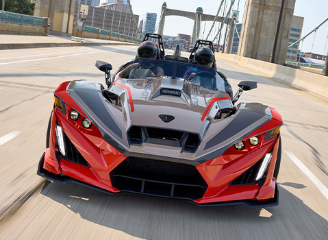 Red and gray Polaris Slingshot autocycle driving across a city bridge with two helmeted riders.