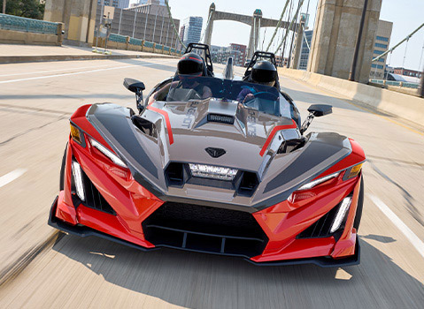 Red and gray Polaris Slingshot autocycle driving across a city bridge with two helmeted riders.