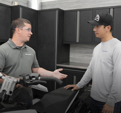 Two men talking next to a Sportsman ATV. The man on the left is a Polaris Product Pro and wears safety glasses and gestures with his hand. The other man on the right wears a Polaris cap and sleeve.