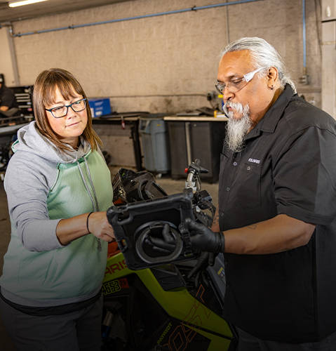 A snowmobile technician holds a tablet displaying service results to rider, with a Polaris Sportsman 570 in the background.