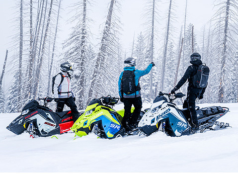 Three people in winter gear stand with three Polaris snowmobiles (red, green, and blue/white) in a snowy forest.
