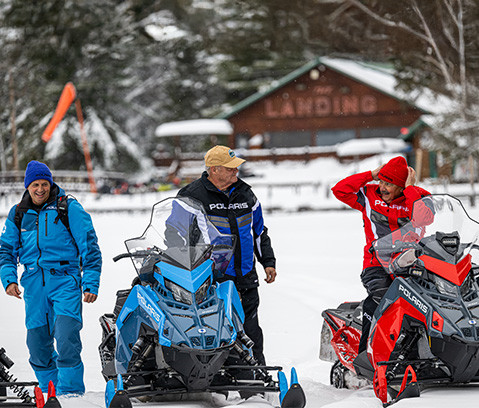 Three men stand with three Polaris snowmobiles (blue, dark blue, and red) in a snowy landscape with a lodge in the background.