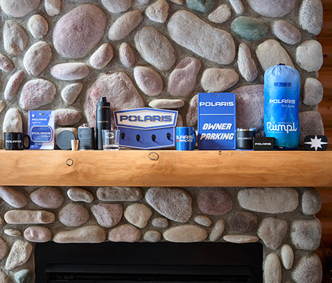Various Polaris-branded merchandise, including mugs, a water bottle, a flask, a key holder, a Polaris Owner Parking sign, a blue sleeping bag, and a small black star-shaped item, are displayed on a wooden mantelpiece above a stone fireplace.